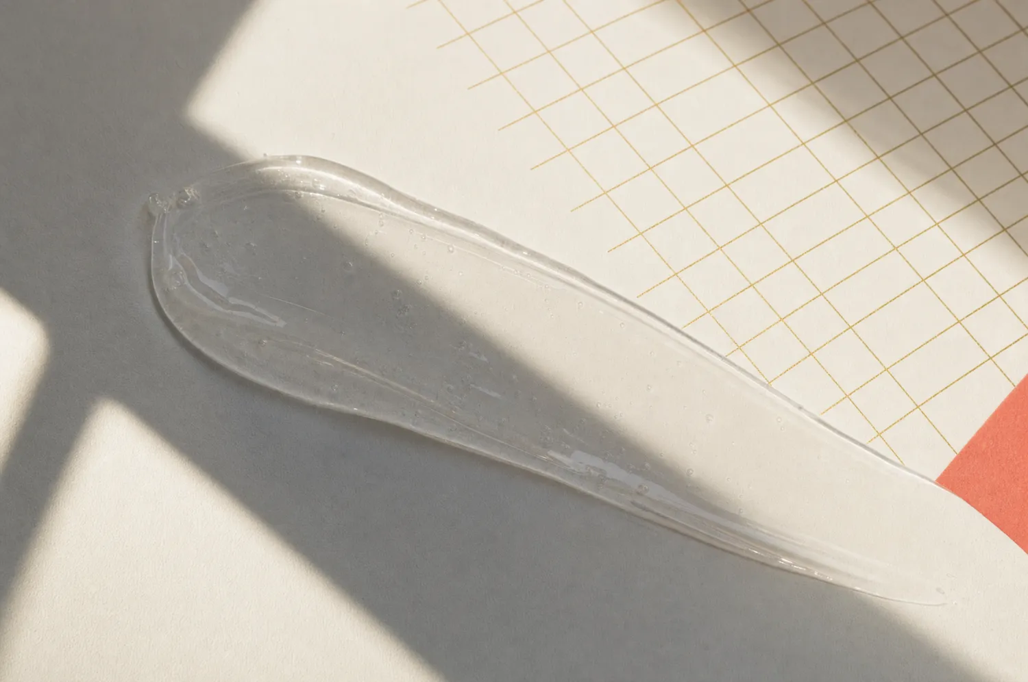 Klarer Sonnenschutz-Gelstrich auf Porzellanfläche, halb im harten Fensterlicht, halb im Schatten, daneben ein feines goldenes Messraster